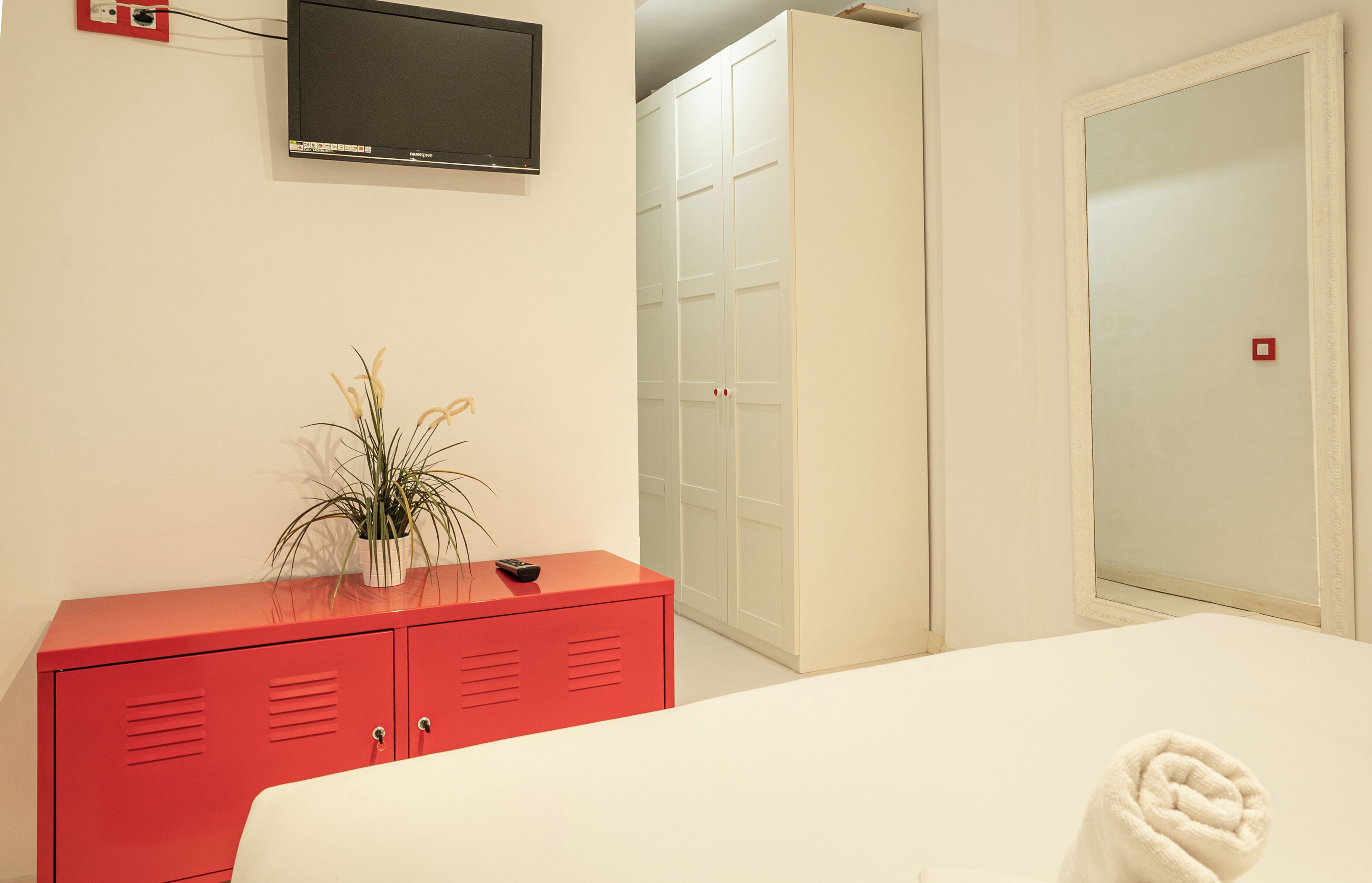 A hotel room with a bed, a red cabinet, a potted plant, a television, and a wardrobe.