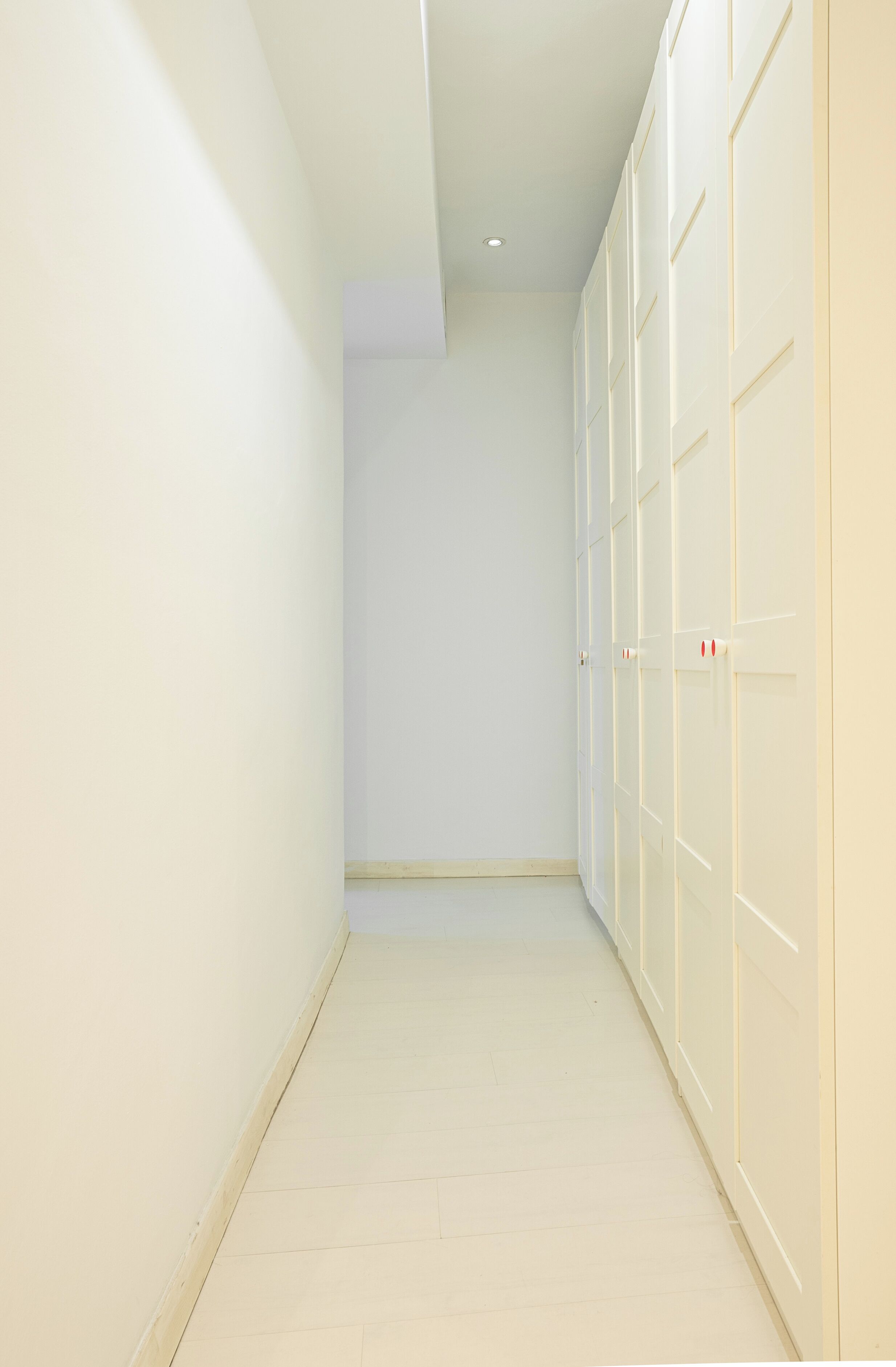 A narrow hallway with white walls and a tiled floor, leading to a set of white closets with visible handles.
