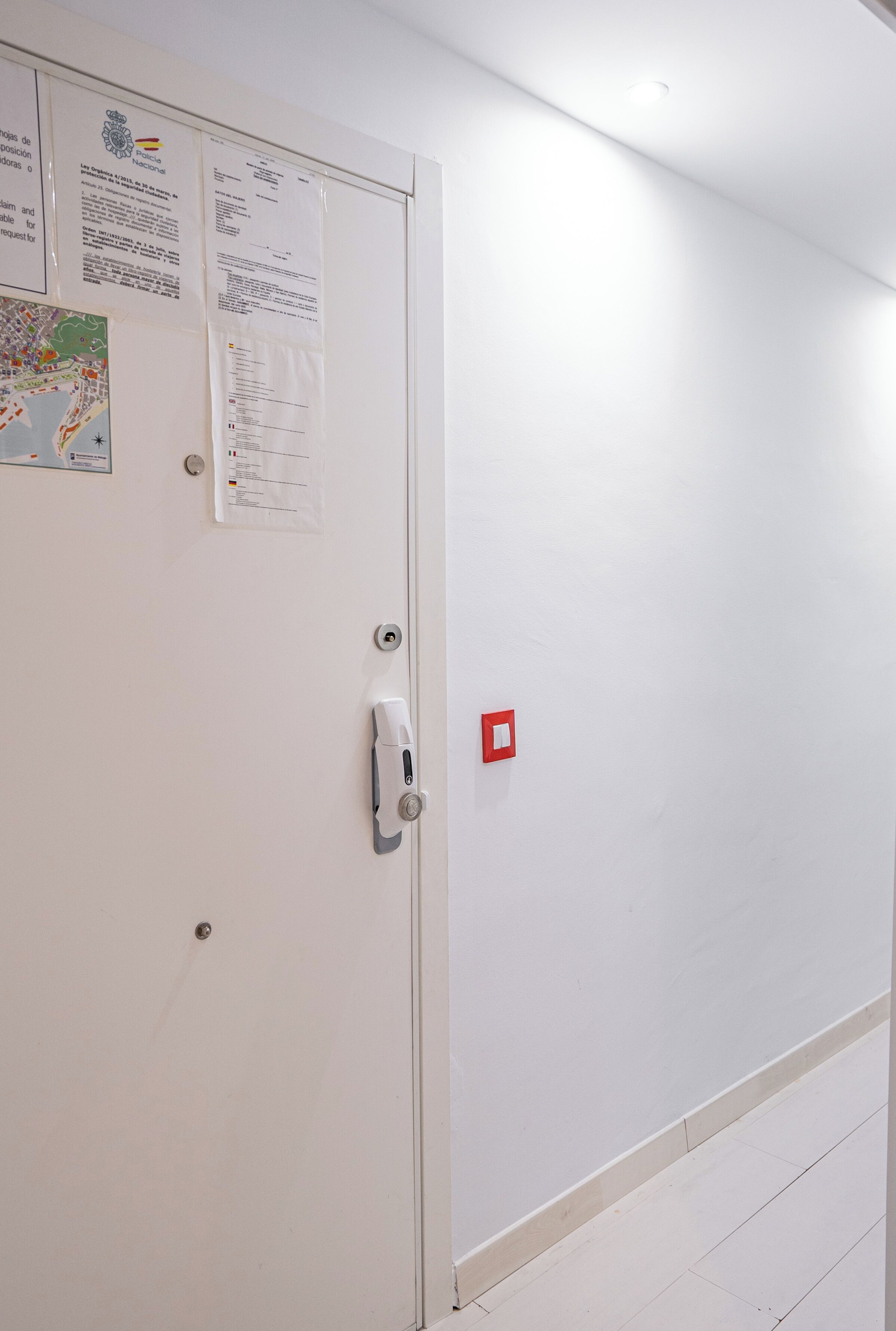 Una puerta blanca con un picaporte, una luz de emergencia roja y varios avisos en la puerta.