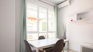 A dining area with a white table and four chairs, a window with curtains, and a shelf with a bottle and a red bowl. - Stay U-nique Apartment Alderete (Málaga)