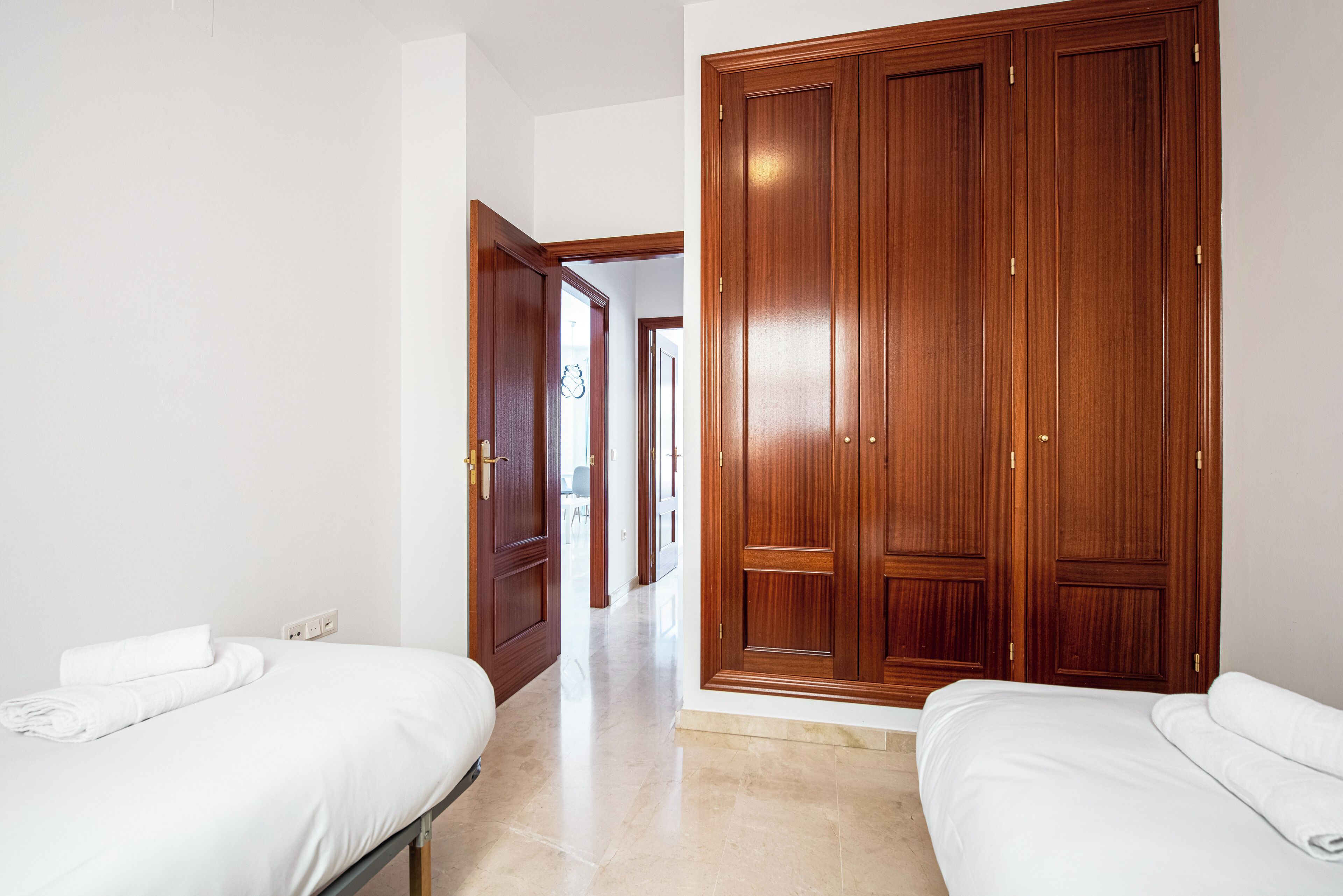 A hotel room with two beds, a wooden wardrobe, and a hallway leading to other rooms.