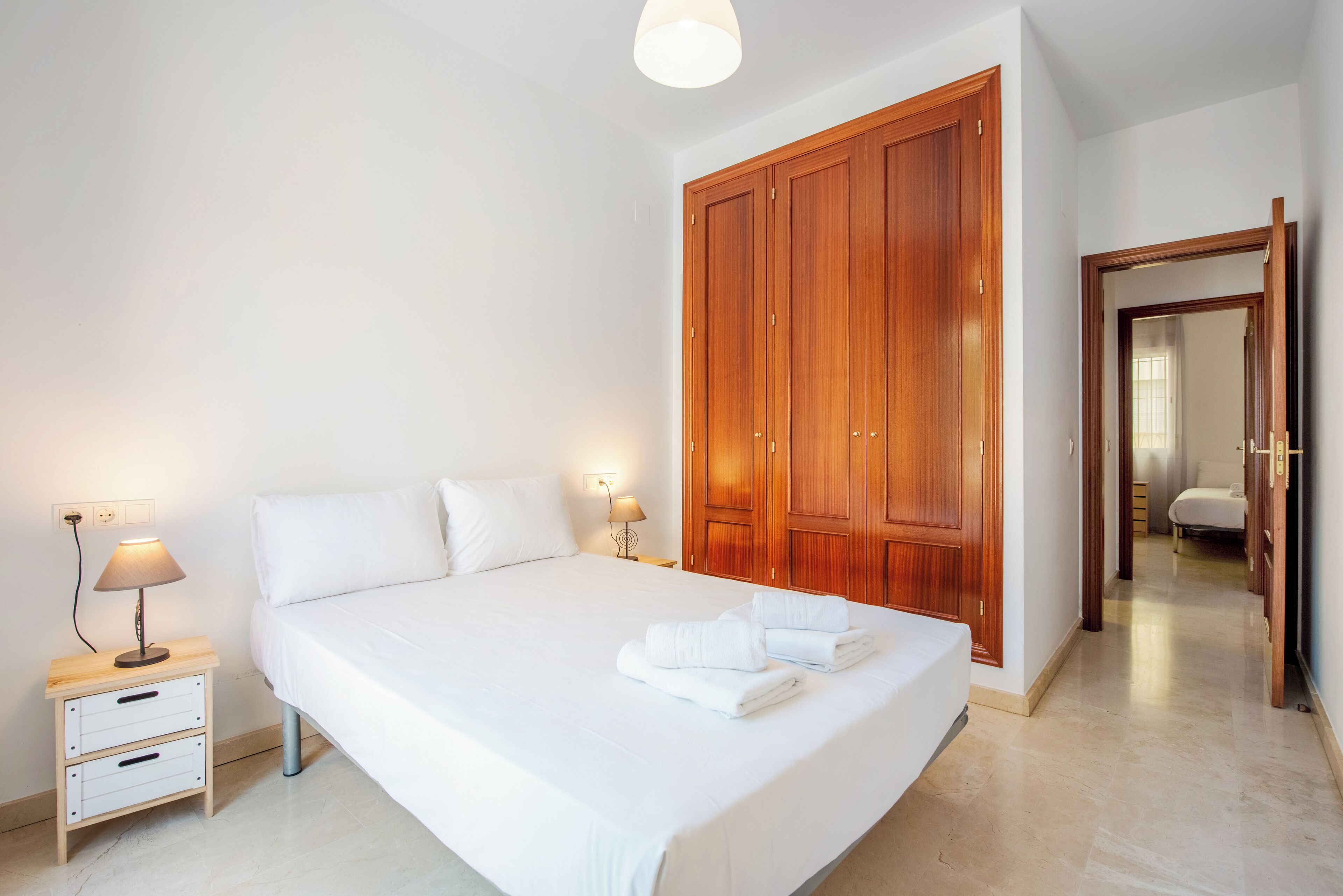 Una cama bien hecha con ropa de cama y toallas blancas, un armario de madera, una mesita de noche con lámpara y un espejo en la habitación contigua.