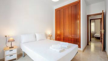 Una cama bien hecha con ropa de cama y toallas blancas, un armario de madera, una mesita de noche con lámpara y un espejo en la habitación contigua.