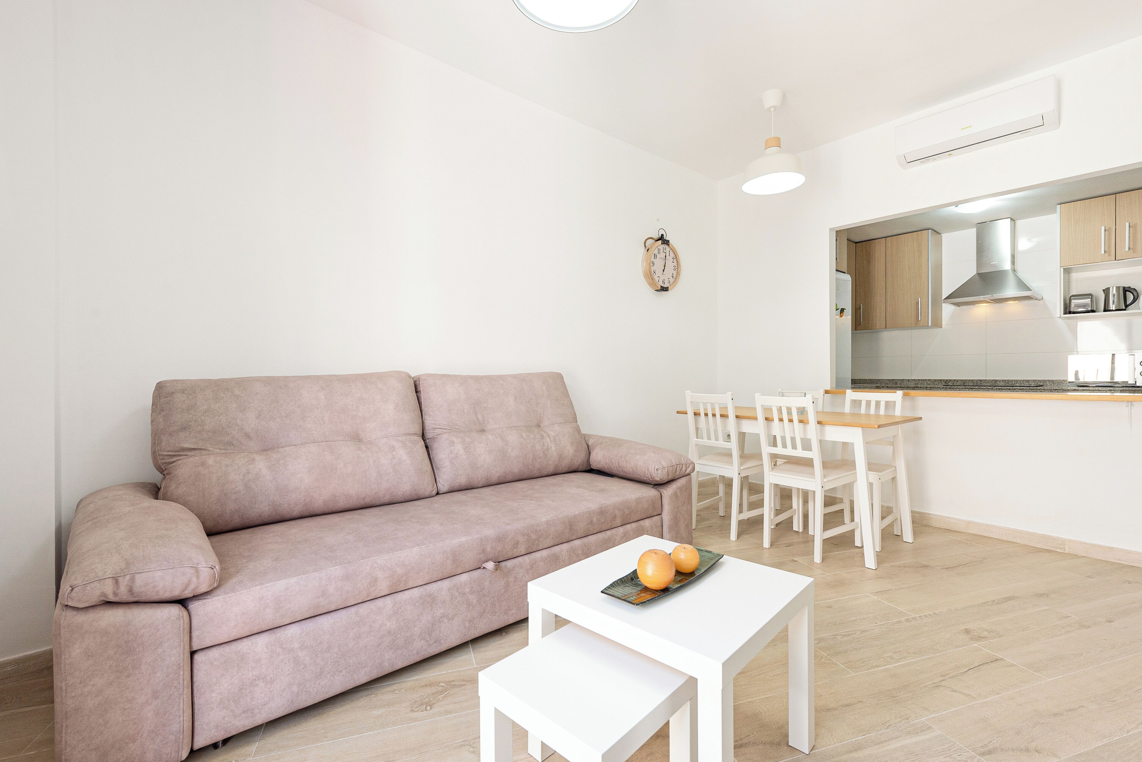 Ein modernes Wohnzimmer mit einer Couch, einem kleinen Tisch mit Orangen und einem Essbereich mit Stühlen, im Hintergrund eine Küche.