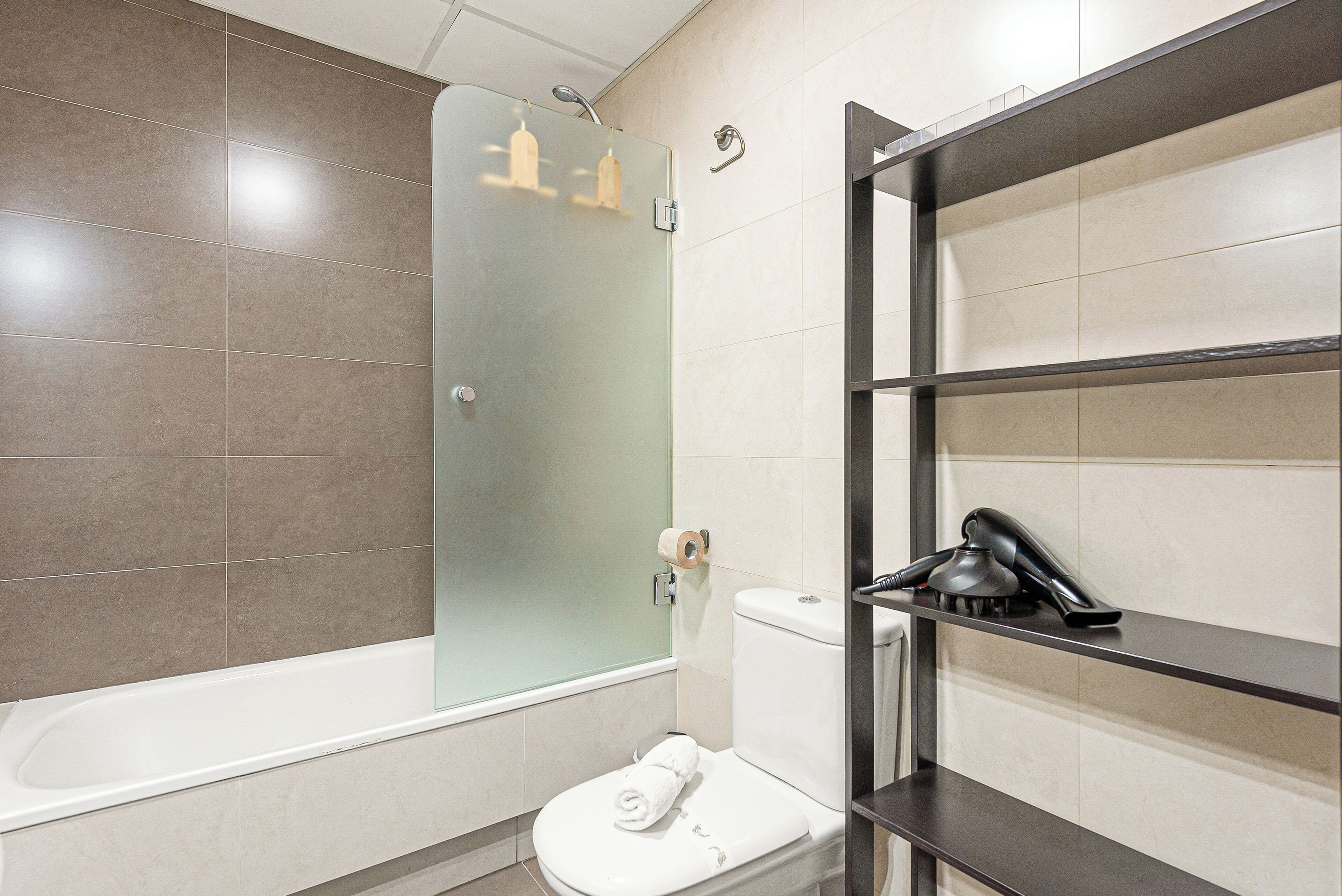 A modern bathroom with a glass-enclosed shower, a white toilet, and a black shelf with toiletries.
