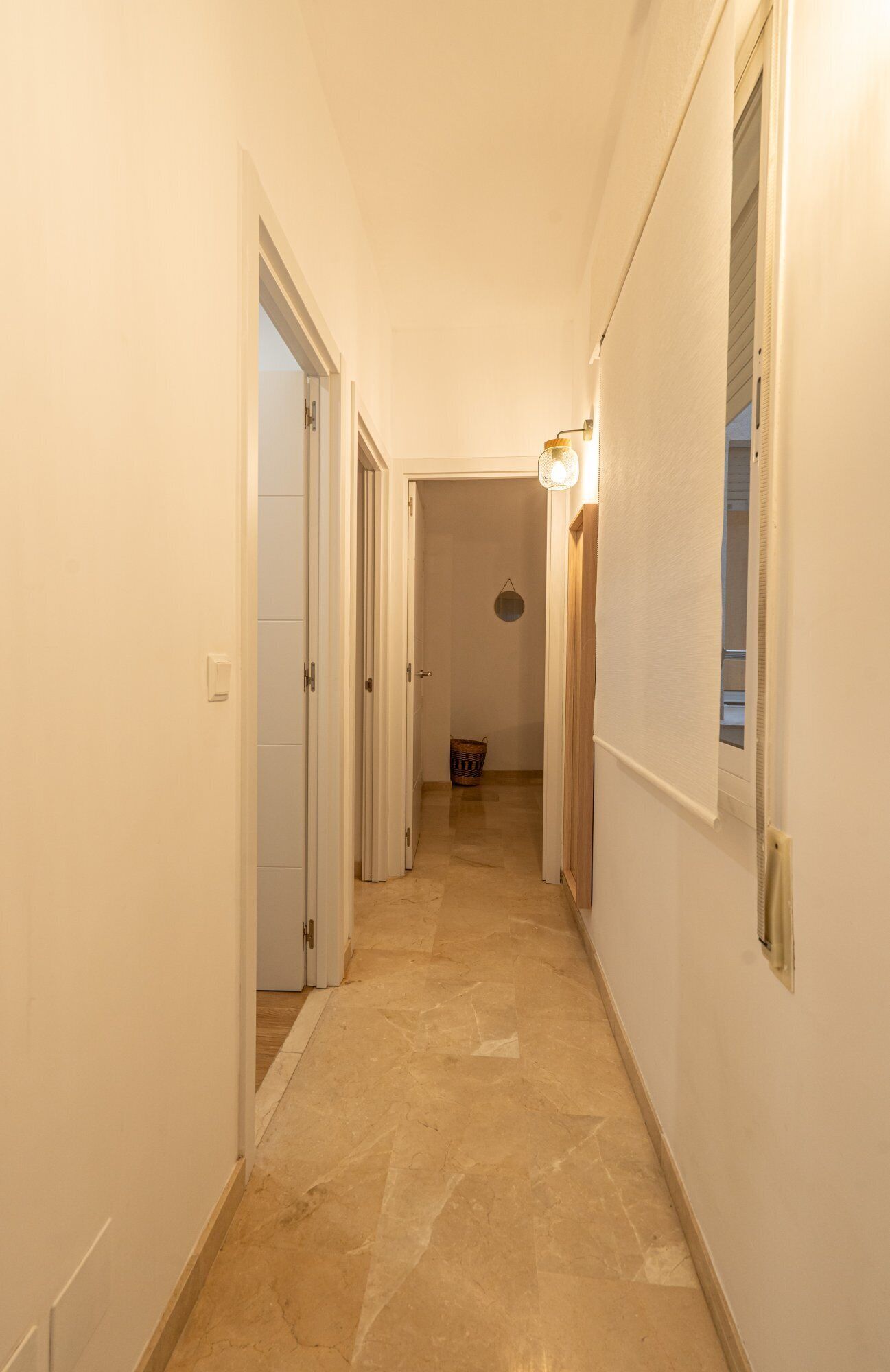 A narrow hallway with white walls and a marble floor. There are doors on both sides and a light fixture on the right wall.
