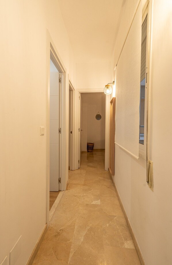 A narrow hallway with white walls and a marble floor. There are doors on both sides and a light fixture on the right wall.