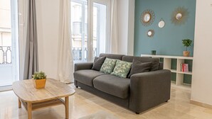 Un salon moderne avec un canapé gris, une table basse en bois et un pouf gris. Il y a un balcon avec vue sur les immeubles.
