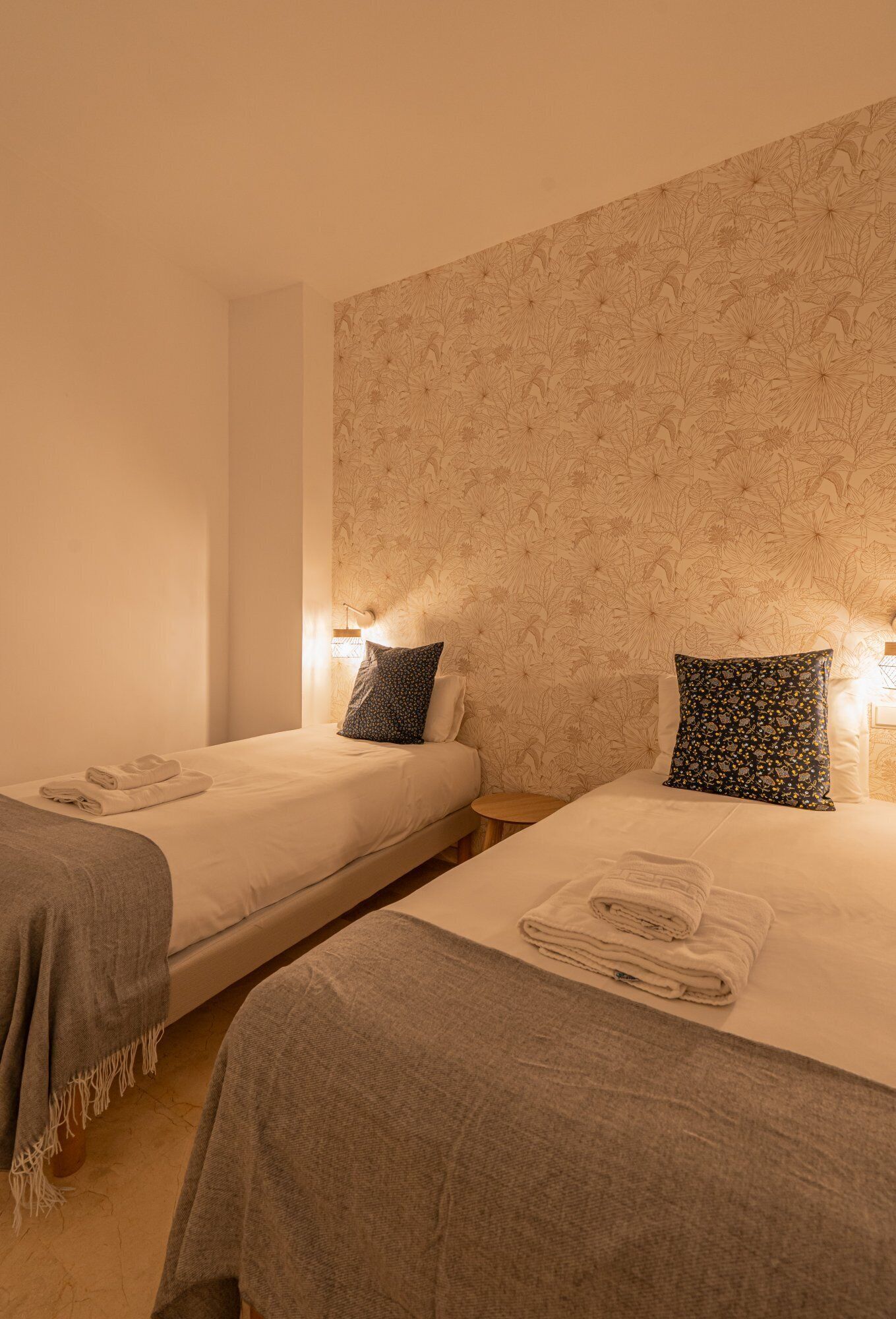 Ein Hotelzimmer mit zwei Einzelbetten, jeweils mit einem Kopfkissen und einem zusammengerollten Handtuch. Das Zimmer hat eine strukturierte Tapete und einen kleinen Nachttisch mit einer Lampe.