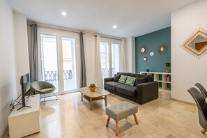 Ein modernes Wohnzimmer mit grauem Sofa, einem kleinen Tisch und einem Fernseher auf einem Schrank. Große Fenster mit Vorhängen, ein Balkon und eine Wand mit dekorativen Spiegeln.