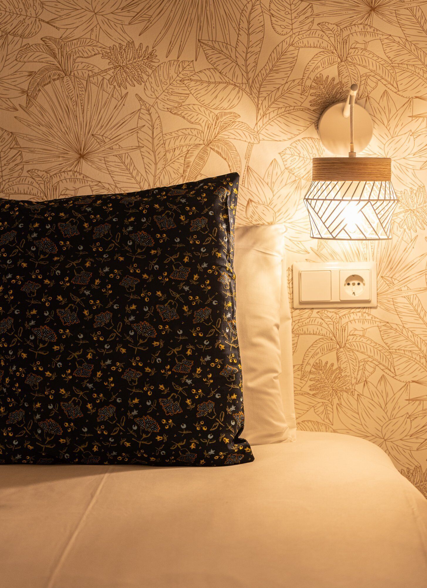 A bed with a dark patterned pillow, a plain white pillow, and a wall-mounted light fixture.