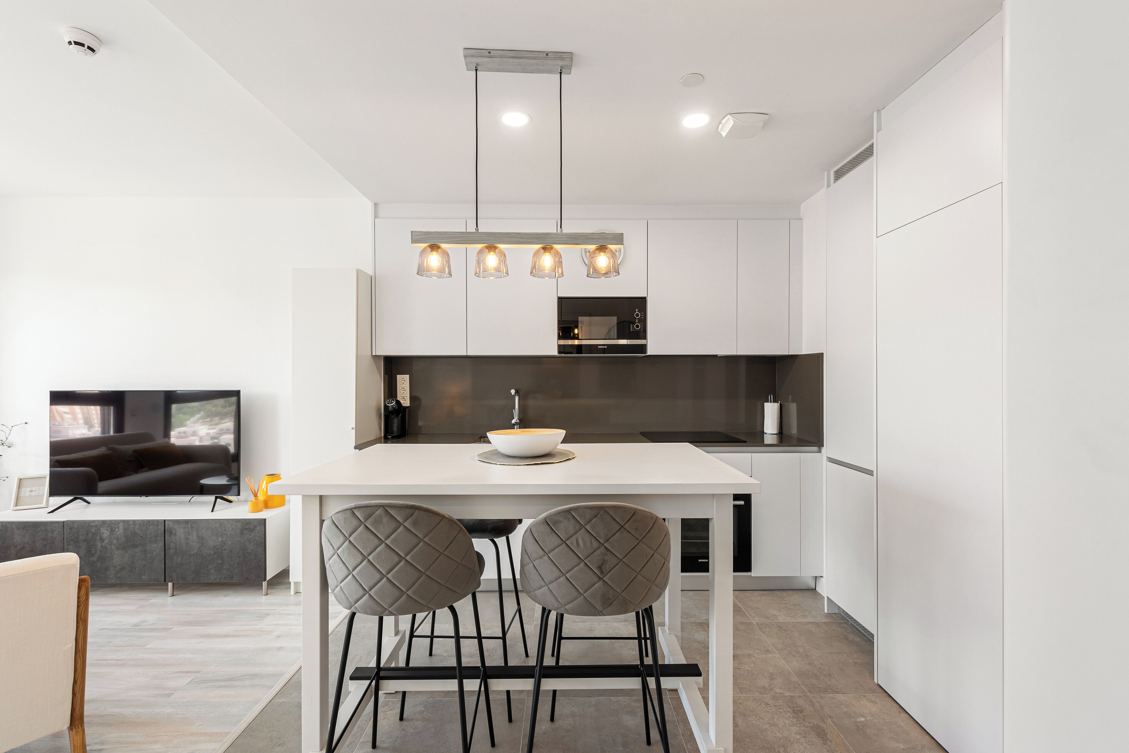 A modern kitchen with a central island, pendant lights, and a flat-screen TV.