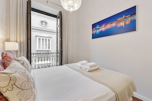A hotel room with a bed, a window with curtains, a painting of a cityscape, and a balcony with a railing.