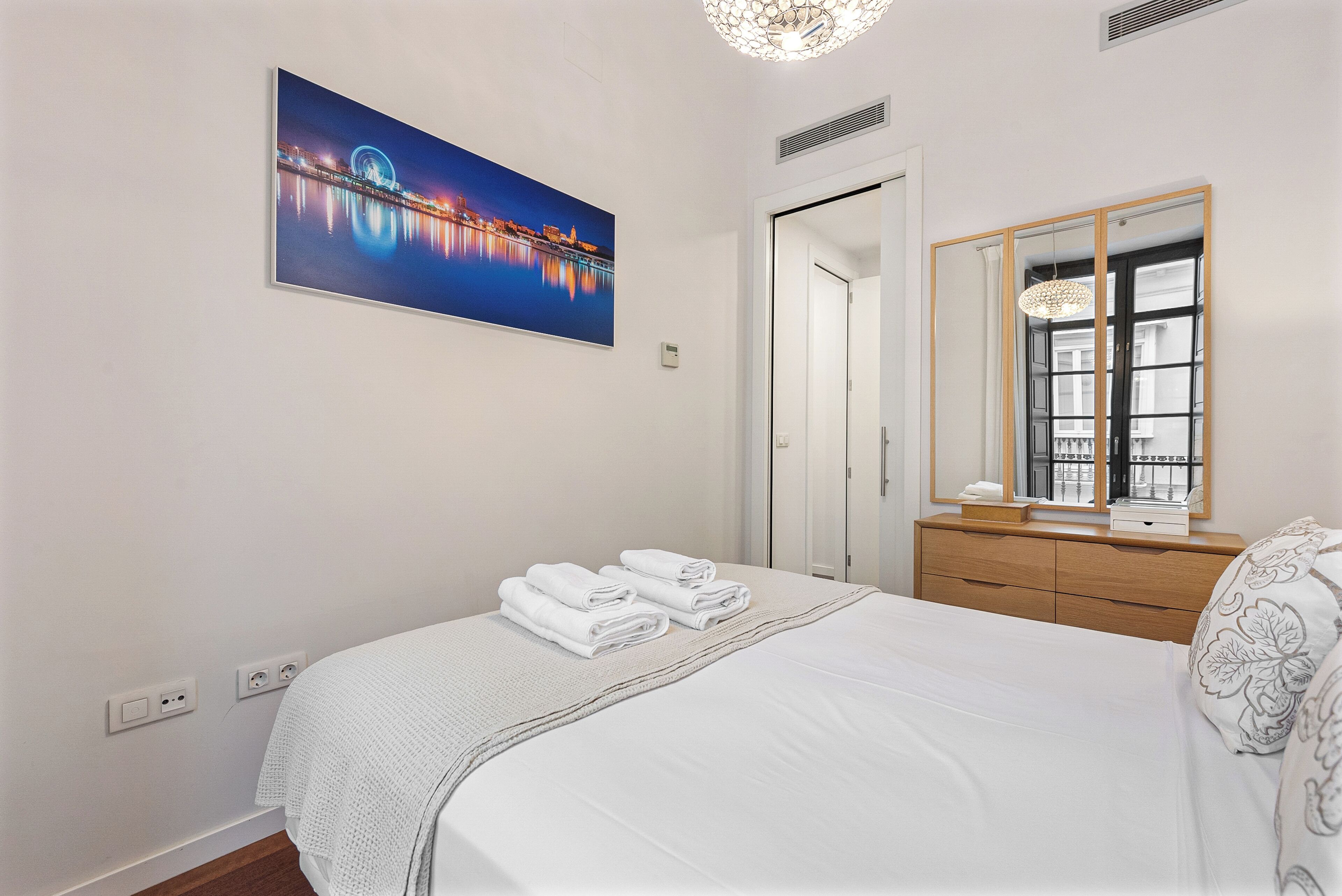 A hotel room with a bed, a nightstand, a mirror, and a painting of a cityscape.