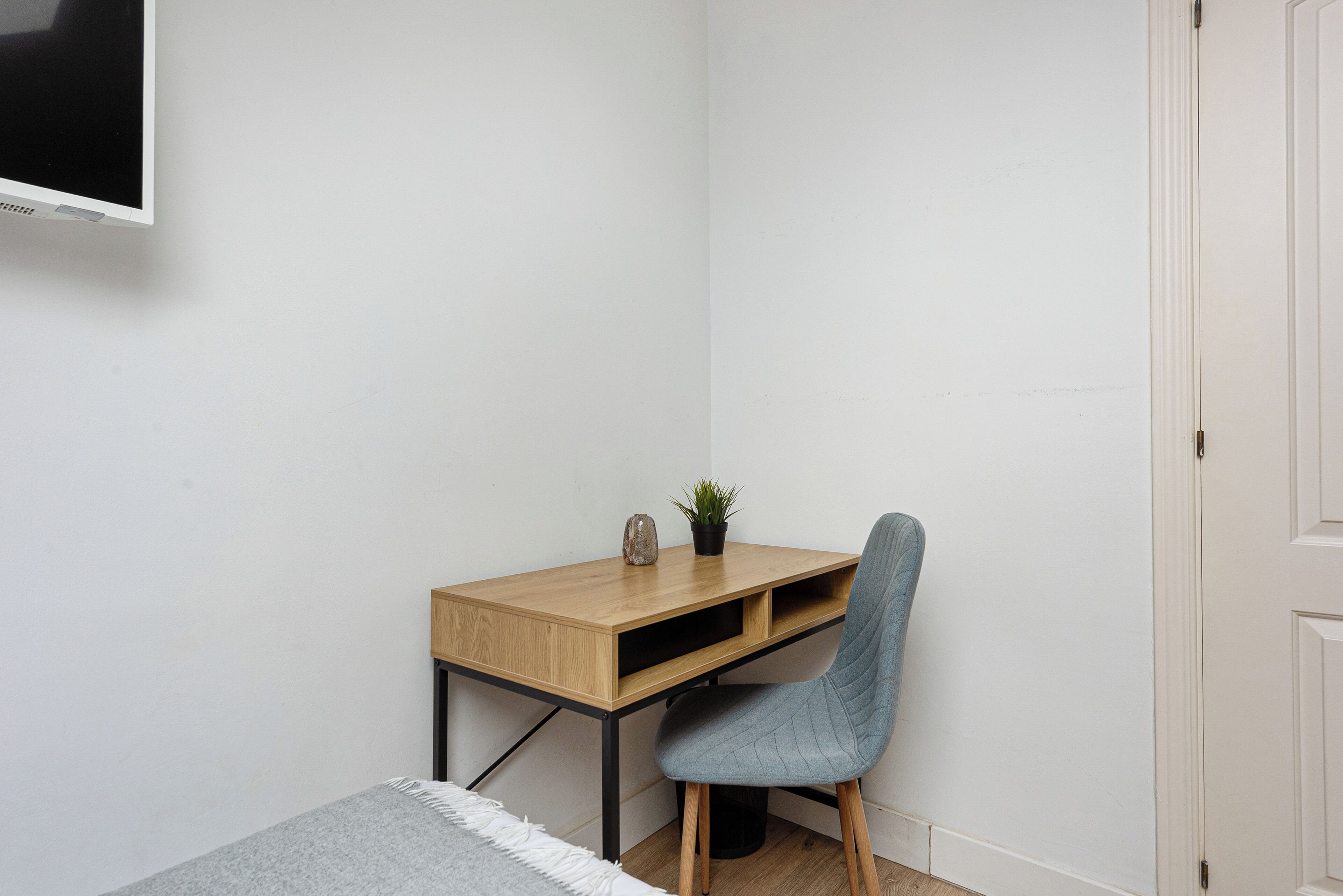 A corner room with a wooden desk, a blue chair, a small potted plant, and a television on the wall.