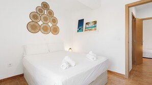 Een net opgemaakt bed met witte lakens en handdoeken, een geweven kunstwerk aan de muur en twee ingelijste foto's van strandtaferelen.