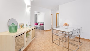 Een moderne woonkamer met een glazen eettafel, een bank met kussens, een houten console tafel met decoratieve objecten, en een gang die naar een andere kamer leidt.