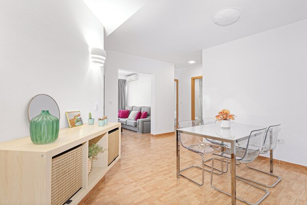 A modern living room with a glass dining table, a sofa with cushions, a wooden console table with decorative items, and a hallway leading to another room. - Stay U-nique Apartment Ferrandiz (Málaga)