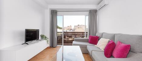 A modern living room with a glass coffee table, grey sofa with pink cushions, a flat-screen TV on a white console, and a view of buildings through large windows.