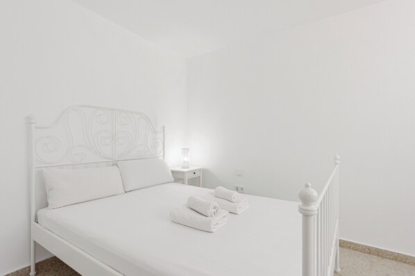 A white bed with a decorative headboard, two rolled towels, a nightstand with a lamp, and a white railing.