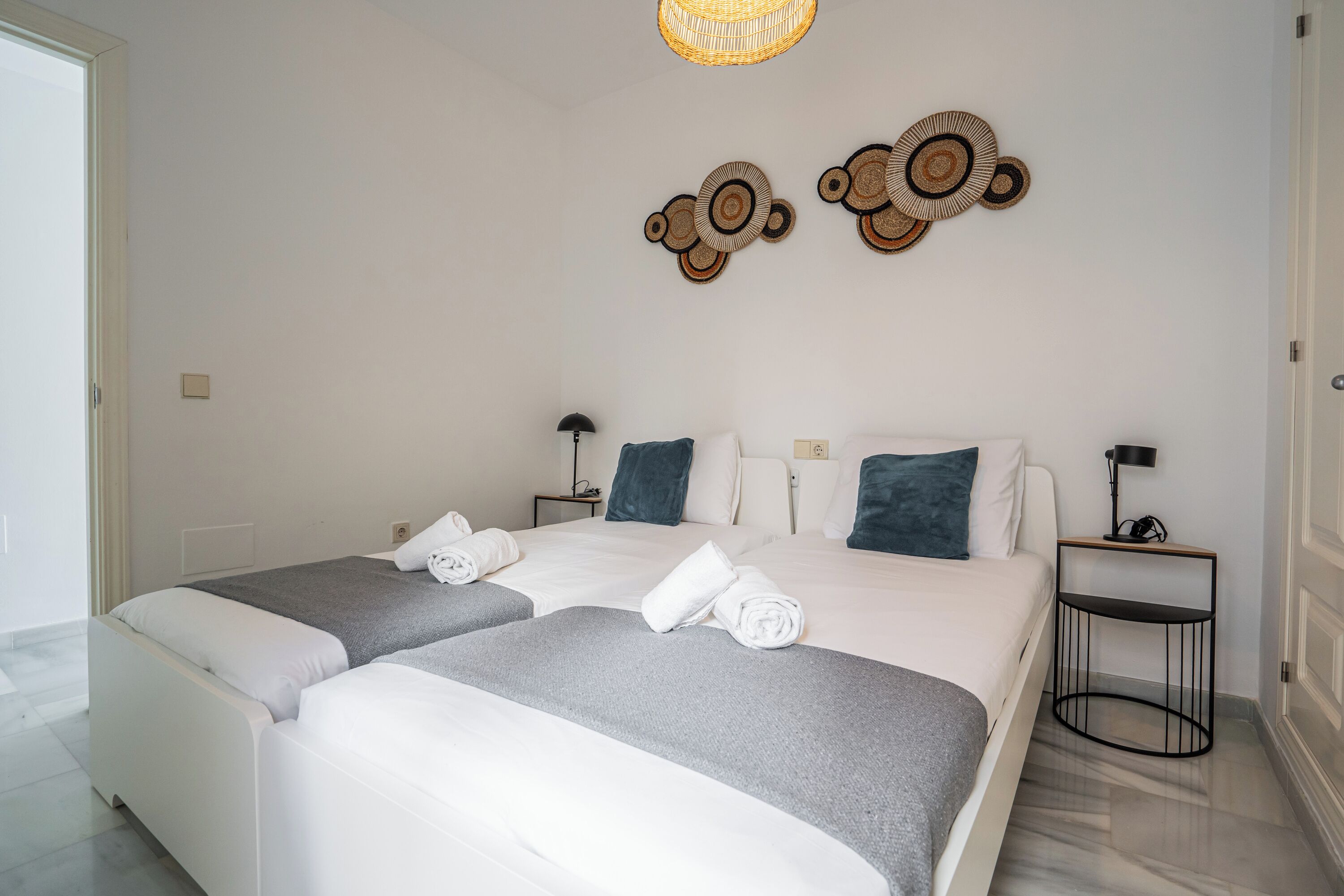 Hotellihuone, jossa on kaksi sänkyä, pieni sivupöytä, seinään kiinnitetty valaisin ja koristeellinen seinätaideteos.