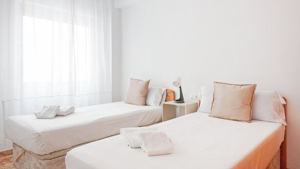 Une chambre d’hôtel avec deux lits simples, une table de chevet avec une lampe et une fenêtre avec des rideaux légers.
