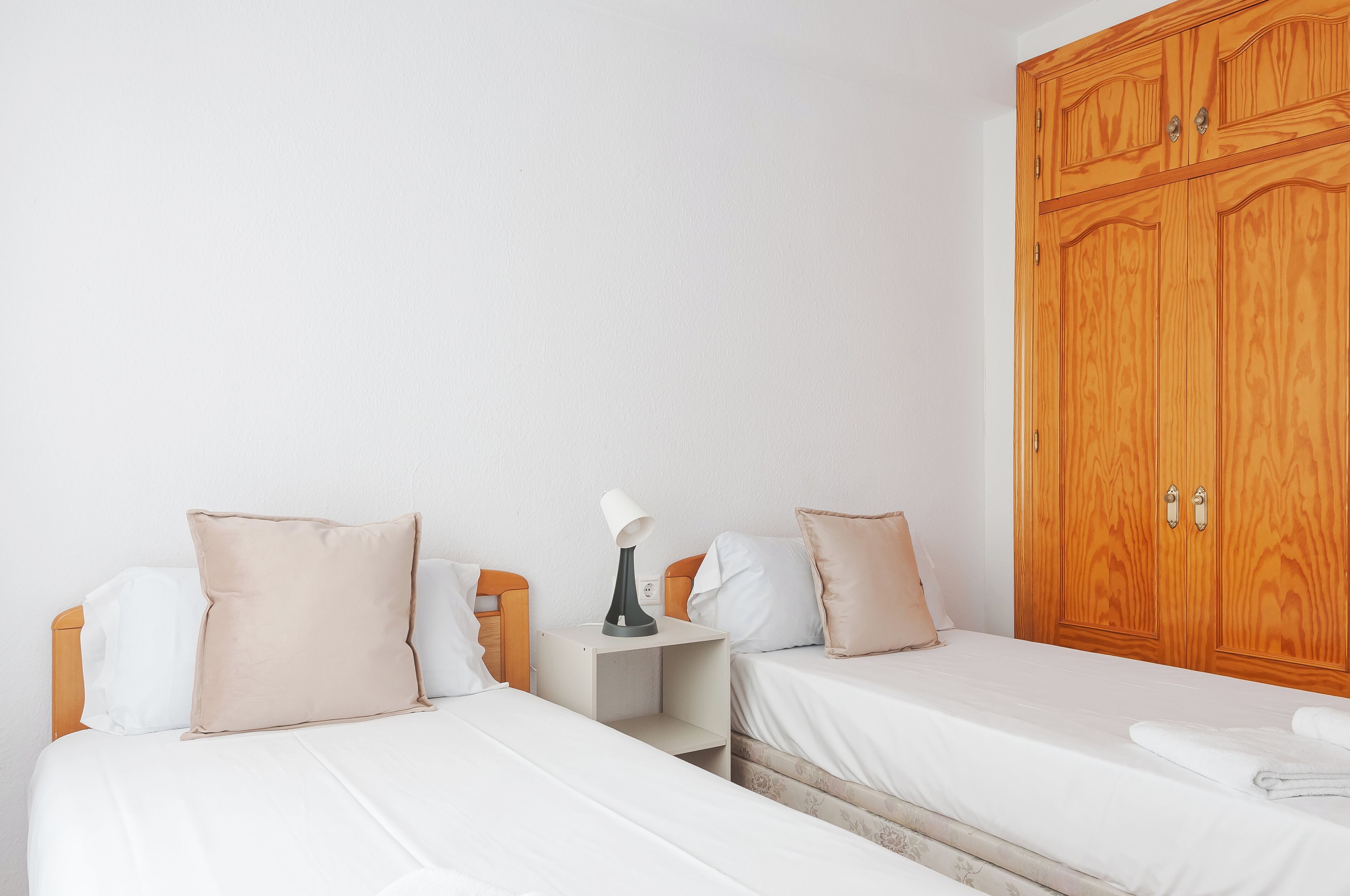 Habitación de hotel con dos camas individuales, un armario de madera, una mesita de noche con lámpara y otra mesita auxiliar.