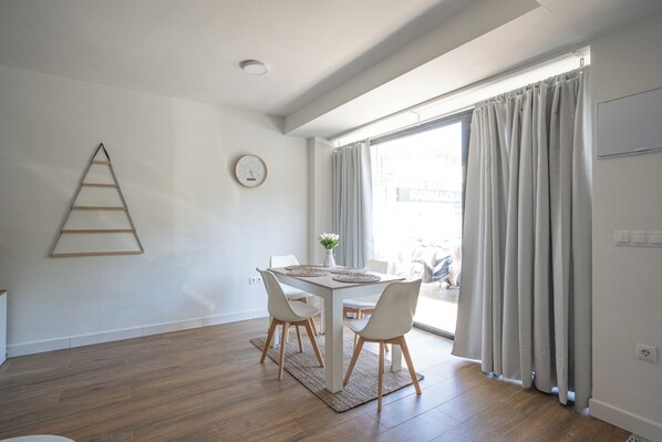 A modern dining area with a wooden table, four chairs, a clock, and a view through large windows with curtains. - Stay U-nique Apartment Pintor Joaquin Sorolla (Málaga)