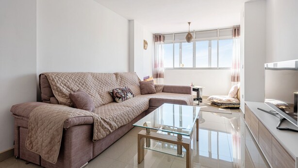 Un salon moderne avec un canapé beige, une table basse en verre et une vue sur le ciel par une fenêtre.