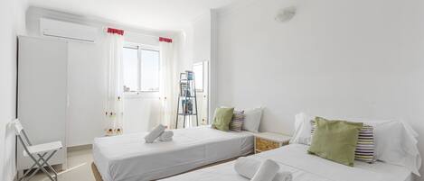 A hotel room with two single beds, a small desk, a chair, a bookshelf, and a window with curtains.