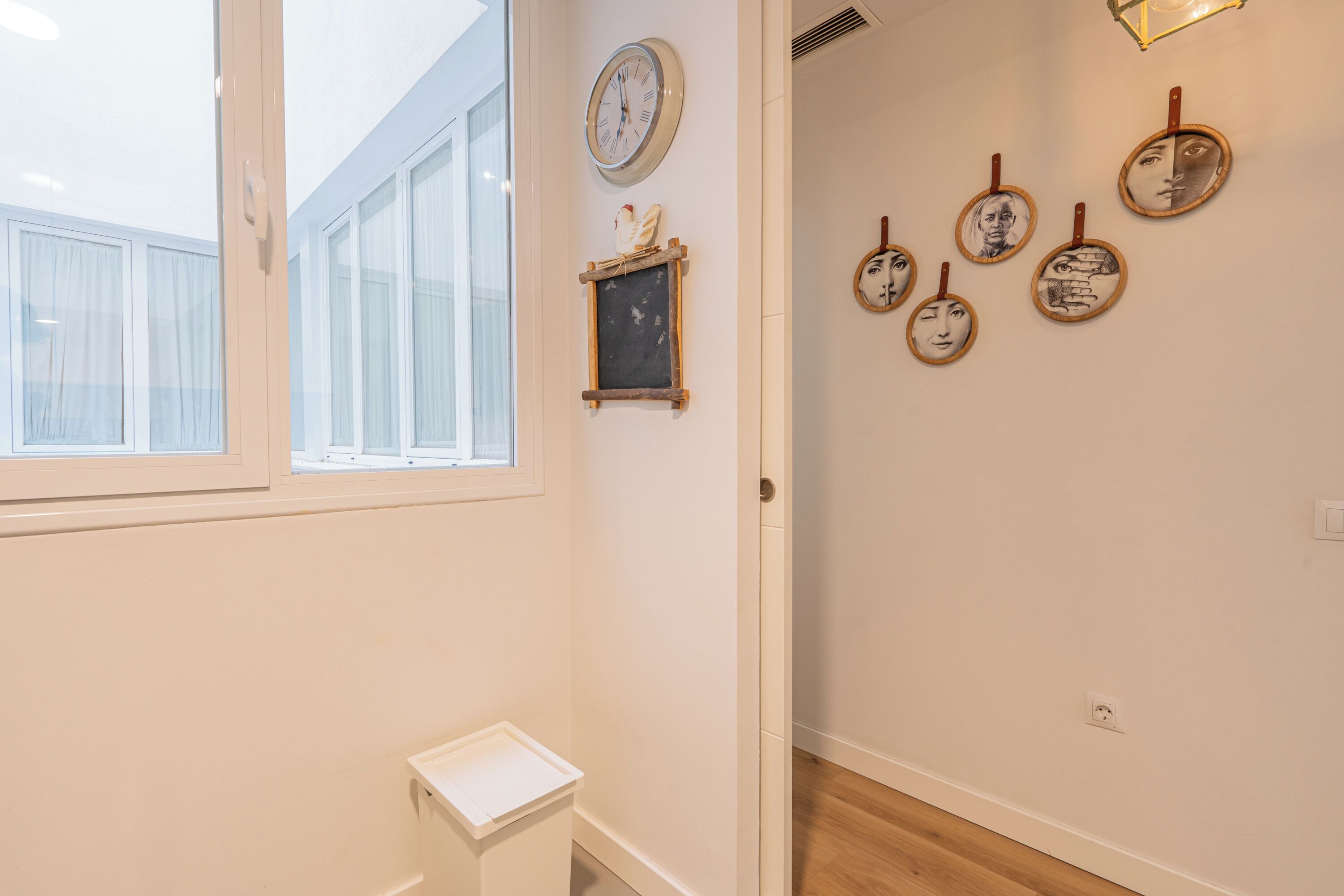 A hallway with a window, a clock, a small shelf with a decorative item, and several framed pictures on the wall.