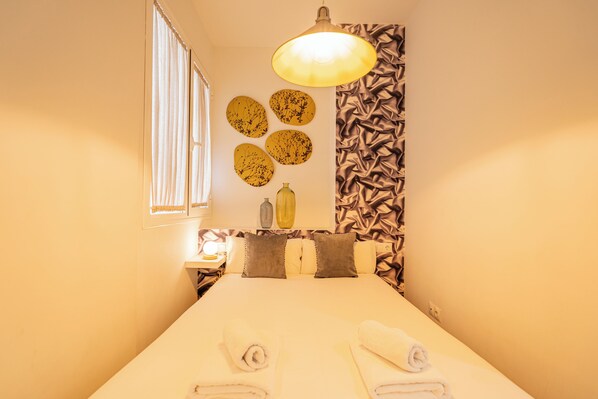 Una cama bien tendida con toallas enrolladas, una lámpara de noche y decoración en la pared, incluyendo platos redondos y un jarrón.
