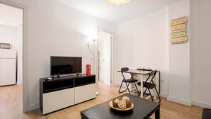 A modern living room with a flat-screen TV, a dining table with chairs, a small cabinet, and a wall-mounted light fixture.