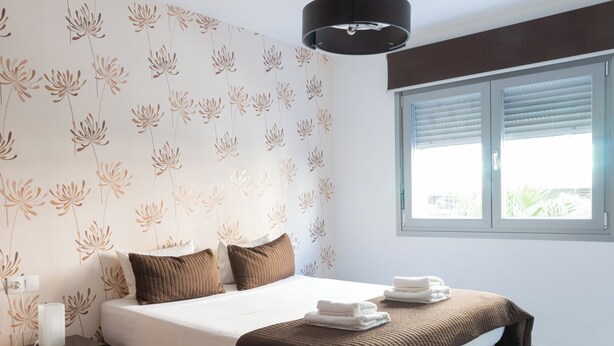 Ein Schlafzimmer mit Bett, Kissen, Nachttisch, Lampe und einem Fenster mit Jalousien.