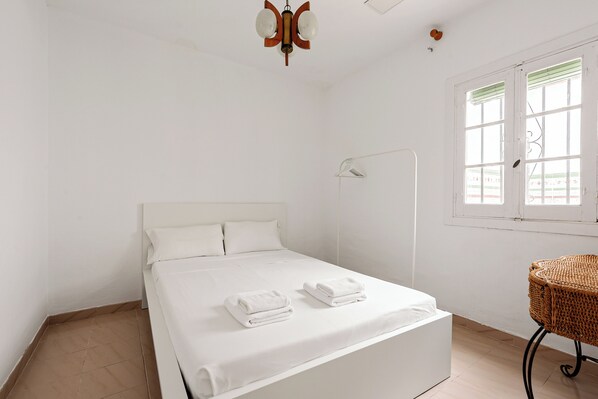 Una habitación limpia y blanca con una cama, dos toallas dobladas, una silla de mimbre y una ventana con persianas.