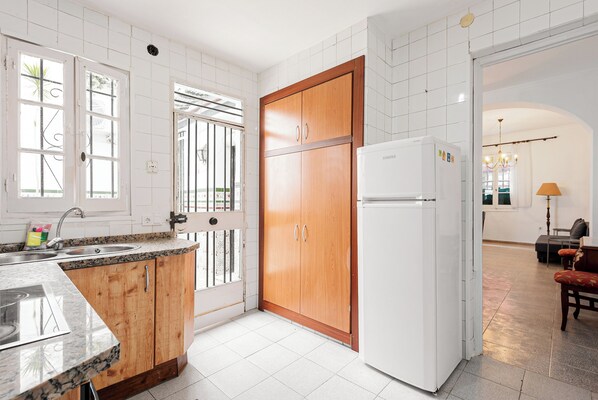 A kitchen with white tiles, wooden cabinets, a refrigerator, and a window with bars.