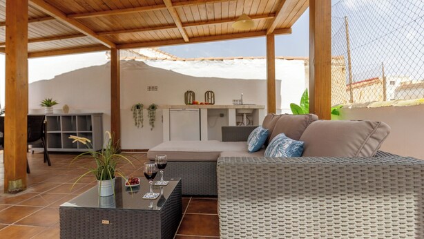 Un espacio exterior techado con sala de mimbre, una mesa con copas de vino y una cocina al fondo.