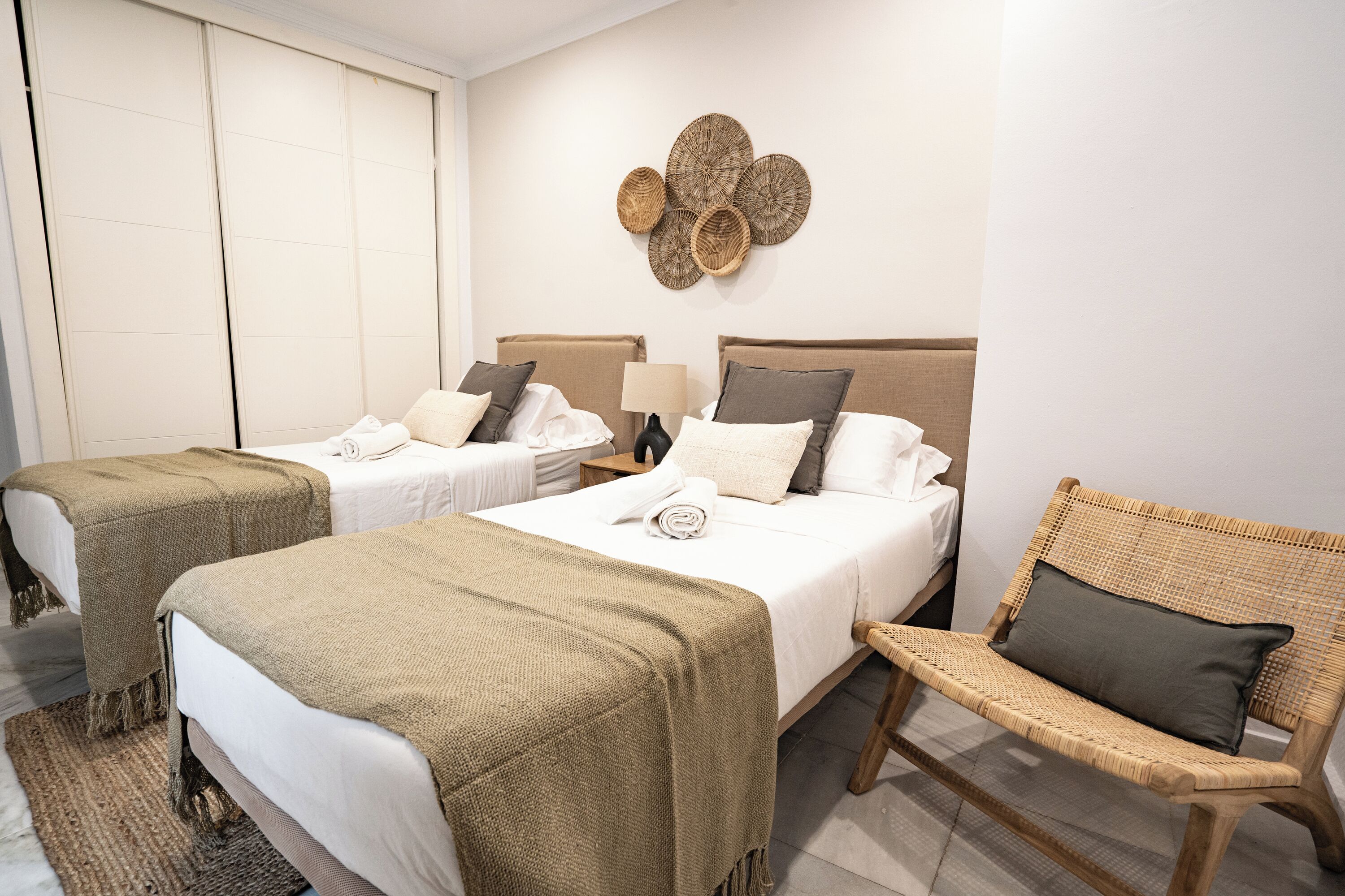 A hotel room with two beds, a wicker chair, and a wall-mounted decorative piece.