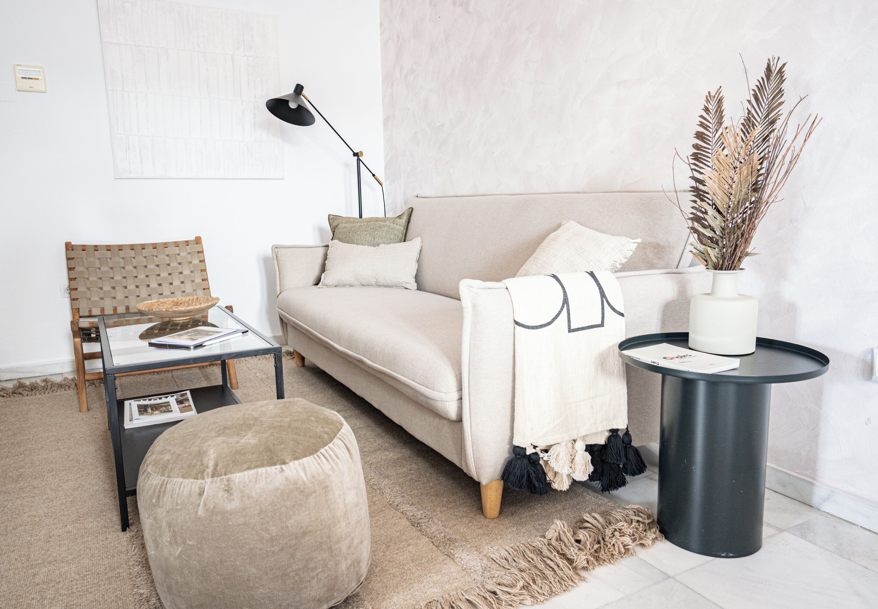 Uma sala de estar moderna com sofá, banqueta, mesa lateral, abajur e planta decorativa.
