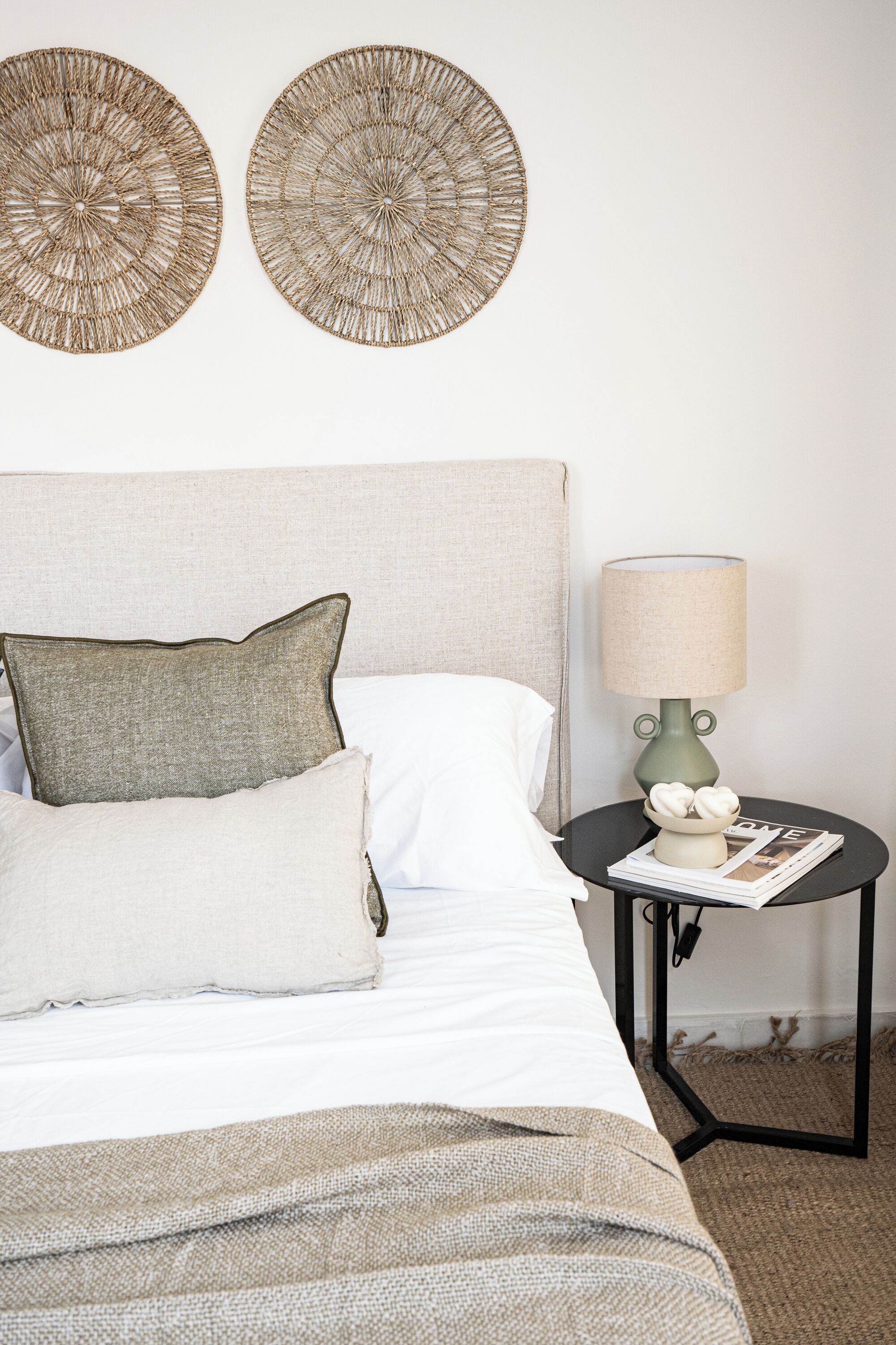 A neatly made bed with a grey headboard, a grey pillow, and a white pillow. There is a round table with a lamp and a magazine on it. Two circular wall decorations are above the bed.