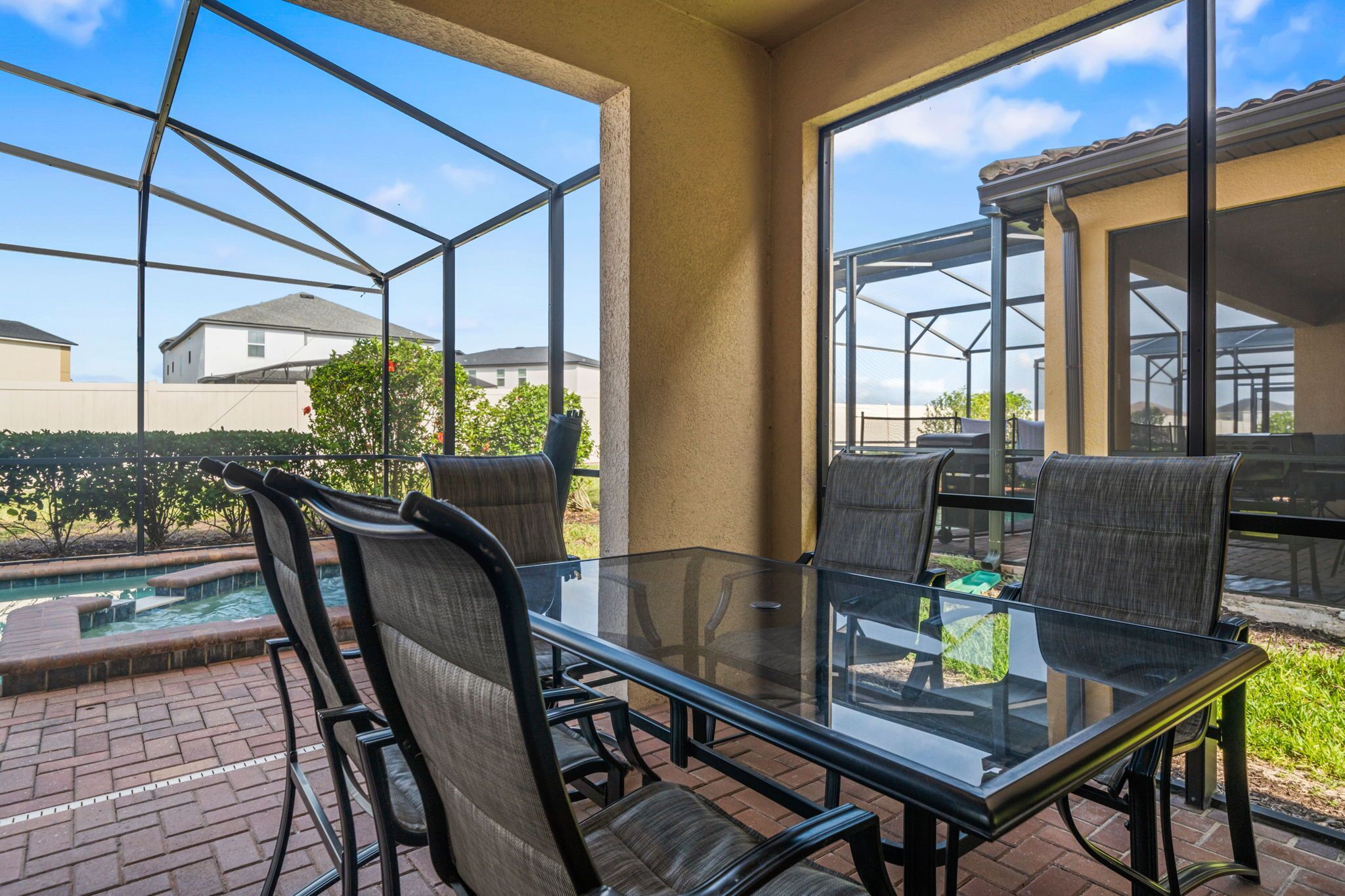 An outdoor patio with a glass table and chairs, a swimming pool, and a view of neighboring houses.