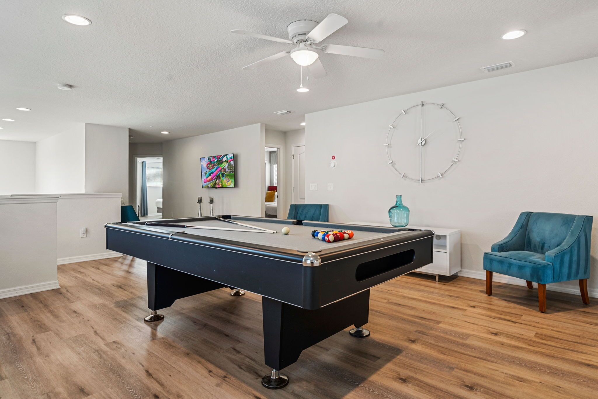 A pool table in a room with wooden flooring, a ceiling fan, and a clock on the wall.