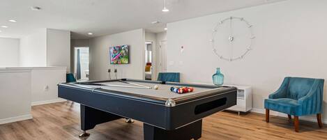 A pool table in a room with wooden flooring, a ceiling fan, and a clock on the wall.