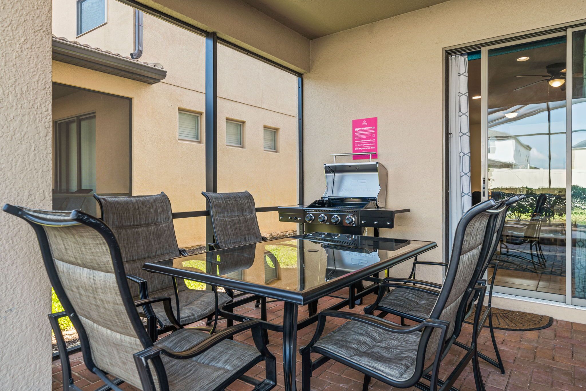 An outdoor patio with a glass table and four chairs, a grill, and a sliding glass door leading to a garden.