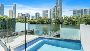 A modern hotel pool with a city view, surrounded by greenery and a calm water body.
