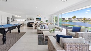A modern living room with a pool table, comfortable seating, a dining area, and a large window overlooking a body of water. - Sanctuary Waterfront – Coastal Elegance in the Heart of Broadbeach Waters (Gold Coast)