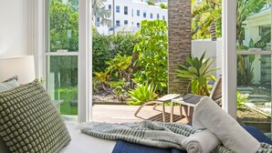 A bedroom with a large window, a bed with a blue quilt, and a view of a garden and buildings outside. - Sanctuary Waterfront – Coastal Elegance in the Heart of Broadbeach Waters (Gold Coast)