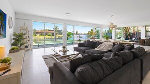 A spacious living area with a large gray sofa, a coffee table, and a view of a body of water and greenery. - Sanctuary Waterfront – Coastal Elegance in the Heart of Broadbeach Waters (Gold Coast)