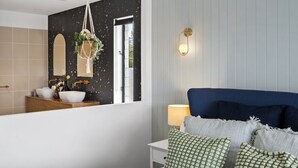 Een slaapkamer met een bed, kussens, een nachtkastje, een lamp, een raam met uitzicht en een badkamer met een wastafel en spiegel.