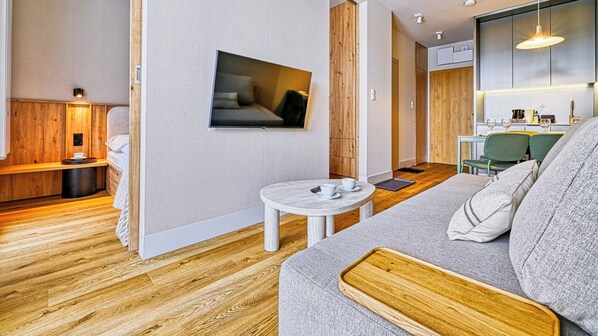 Una sala de estar moderna con piso de madera, un sofá gris, una mesita blanca con tazas y un control remoto, y un televisor montado en la pared.